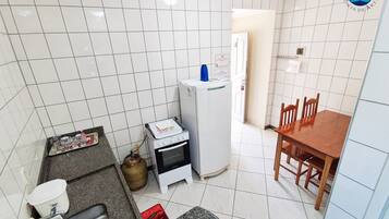 Cozinha compacta com paredes de azulejos brancos, fogão a gás, forno pequeno, geladeira, mesa de jantar com cadeiras e pia com torneira.