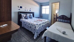 Une chambre d’hôtel avec un lit, un petit lit, un bureau, un ventilateur de plafond, une fenêtre et une salle de bain visible à travers une porte ouverte.