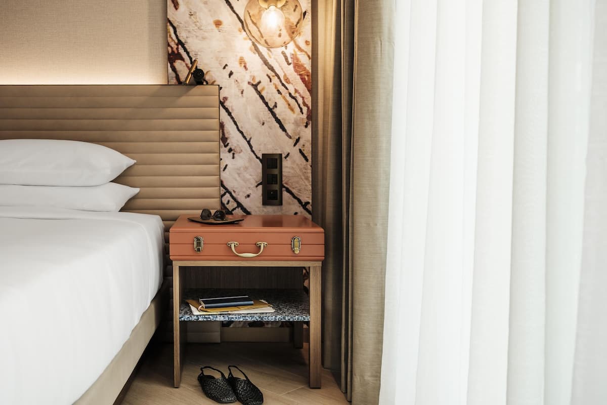 A neatly made bed with a headboard, a wooden nightstand with a lamp, a suitcase, and a shelf with books.