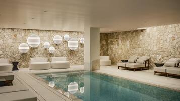 Un área de piscina interior moderna con paredes de piedra, varias zonas de estar y decoraciones geométricas en las paredes.