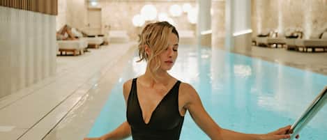 A woman in a black swimsuit standing by a pool with lounge chairs and a spa area in the background.