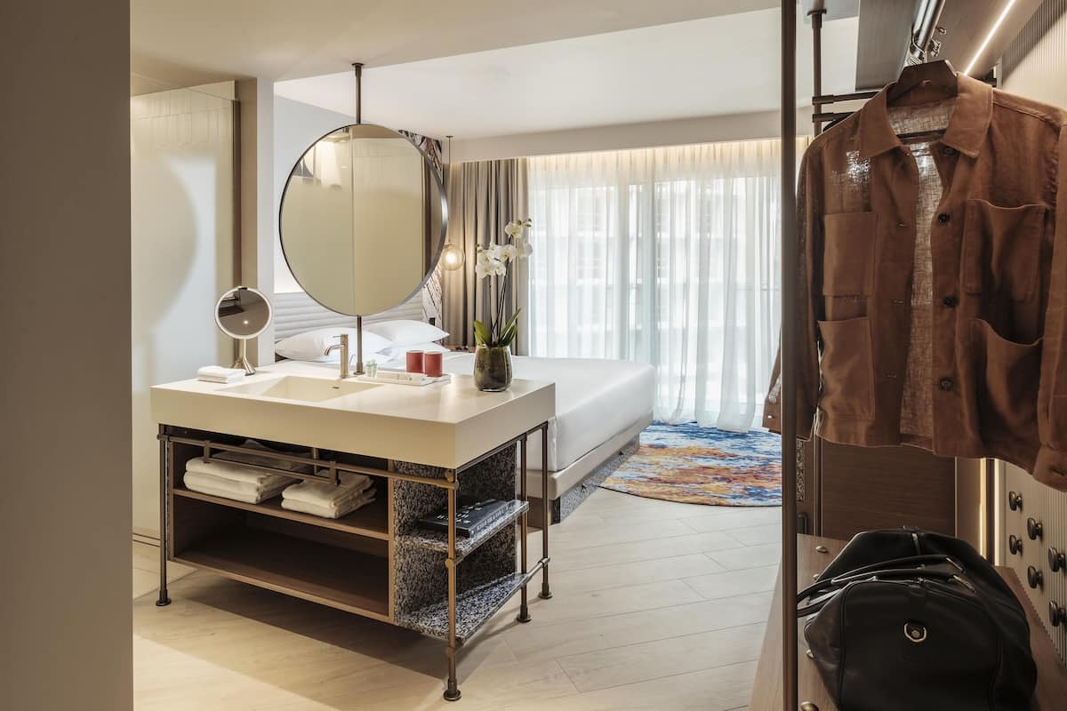 A hotel room with a bed, a round mirror, a vanity with a sink, a robe, and a hanging coat rack.