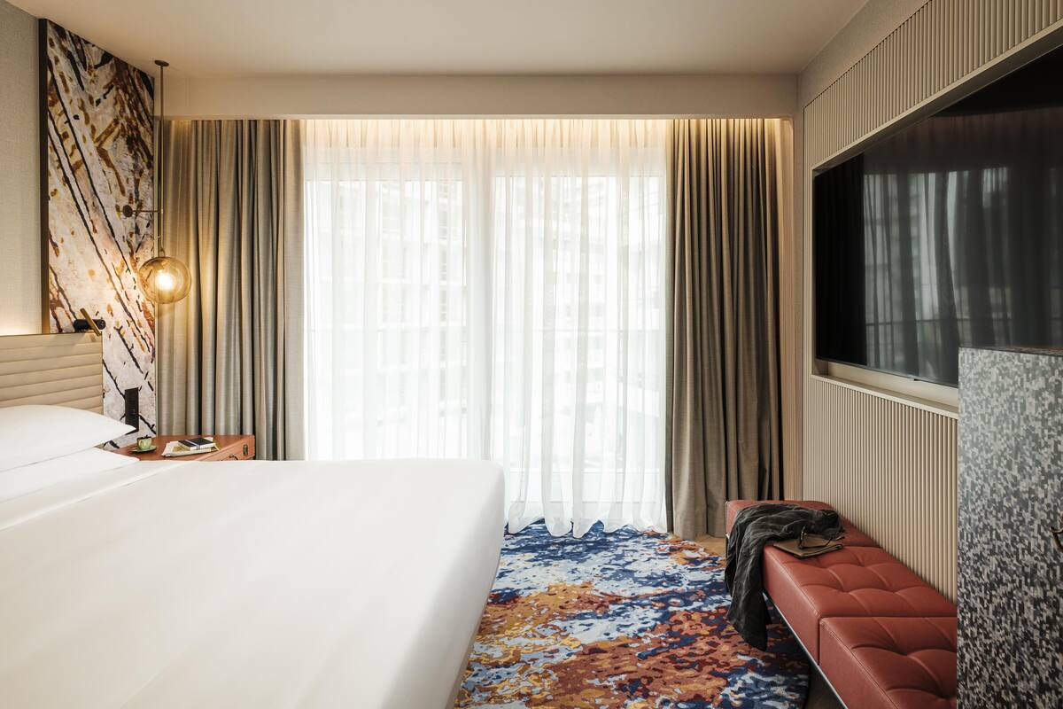 A hotel room with a large bed, a bench, a TV, and a window with curtains.