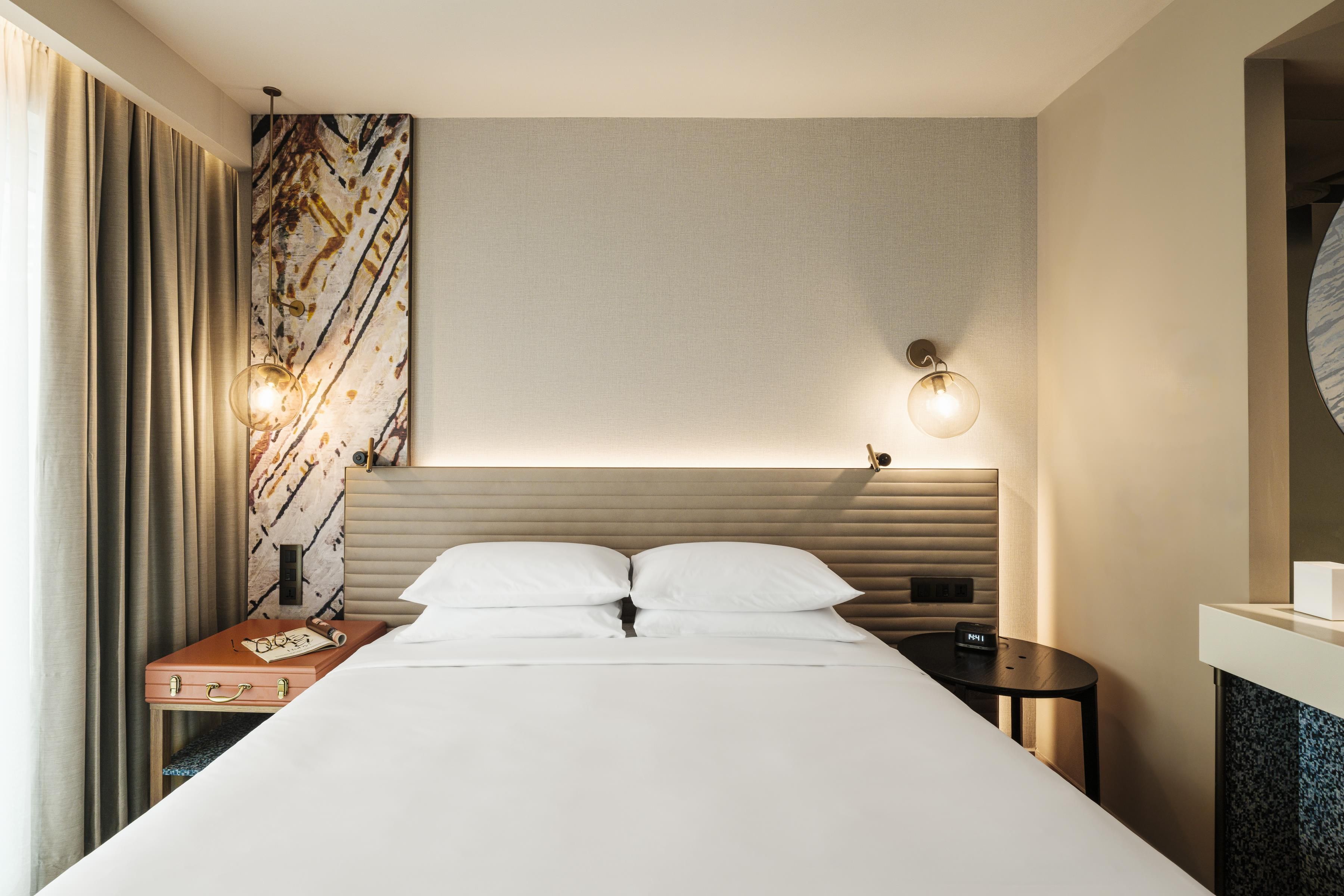 A modern hotel room with a bed, bedside tables, a nightstand, a wall-mounted light fixture, and a decorative wall panel.