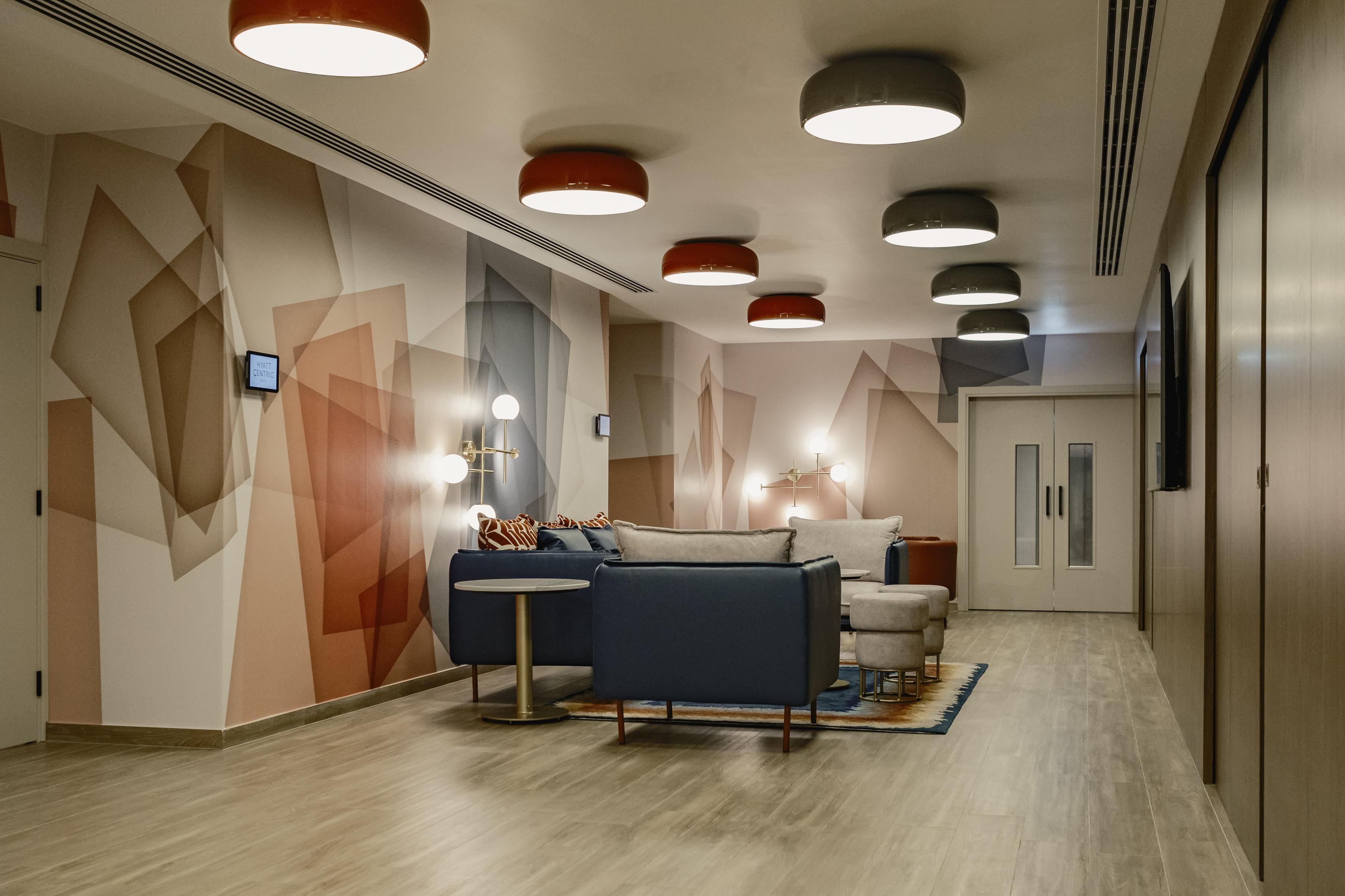 A modern hotel lobby with geometric wall art, a seating area with sofas and a round table, and recessed ceiling lighting.