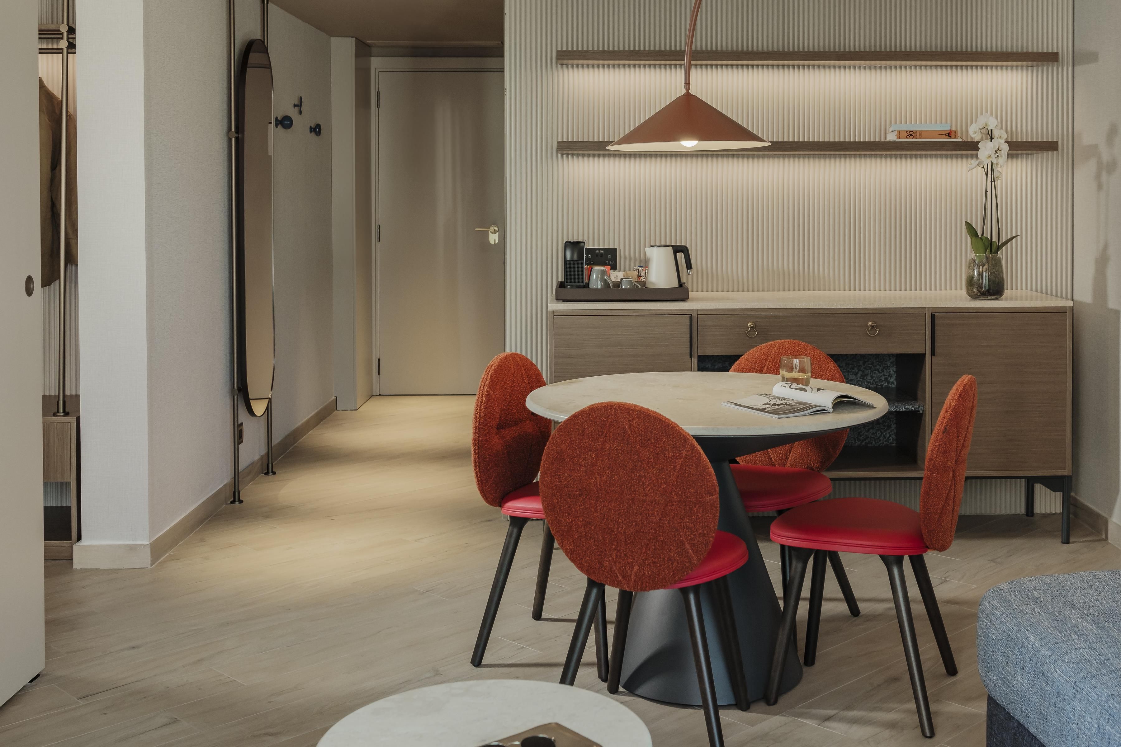 A modern dining area with a round table, red upholstered chairs, a coffee machine, and a vase with an orchid on the wall.