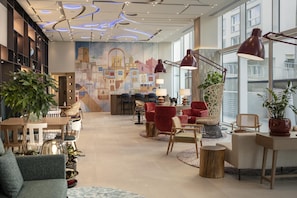 A modern hotel lobby with a large mural, comfortable seating, and a view of the city.