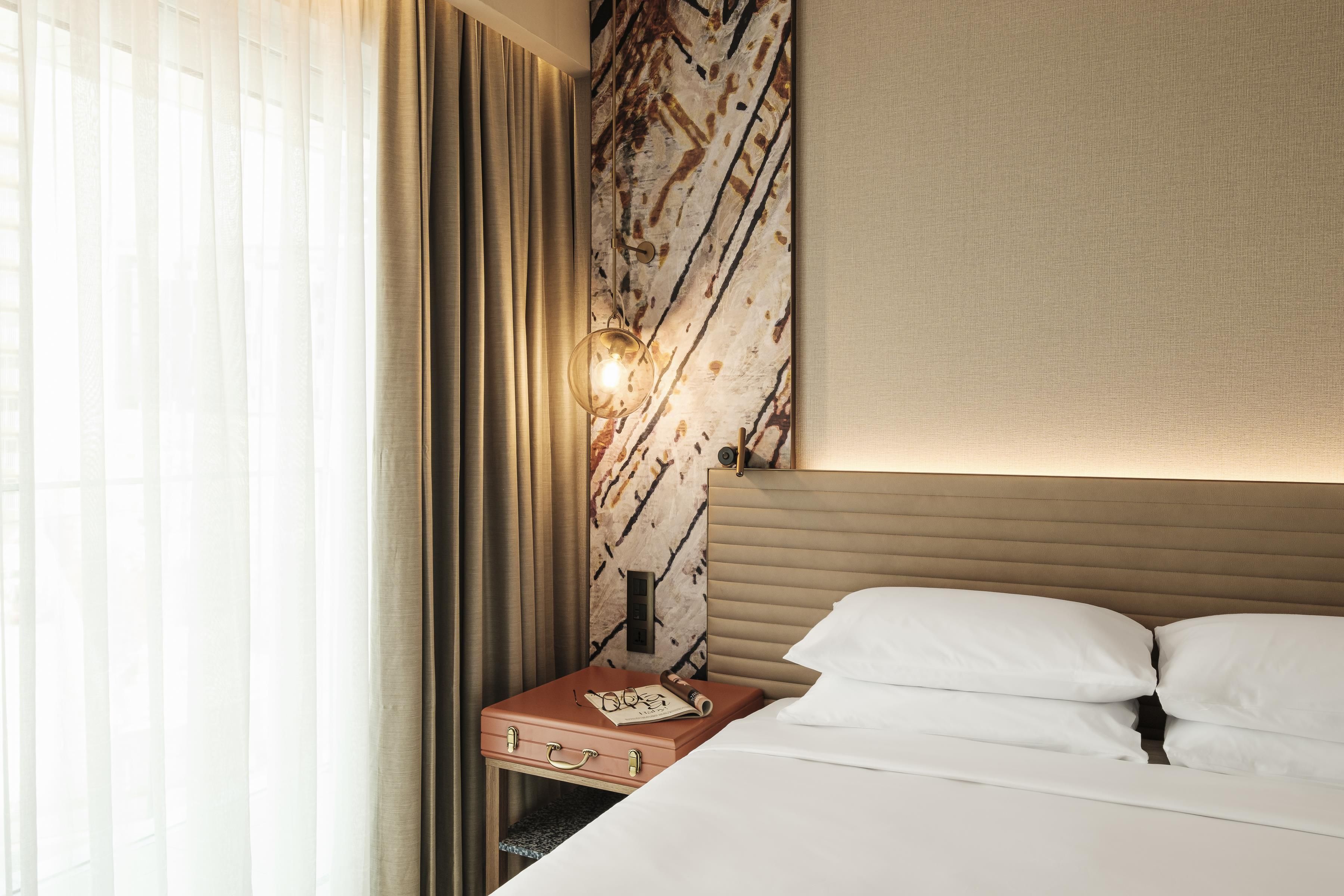 A hotel room with a bed, a nightstand with a lamp, a suitcase, and a wall with a patterned design.