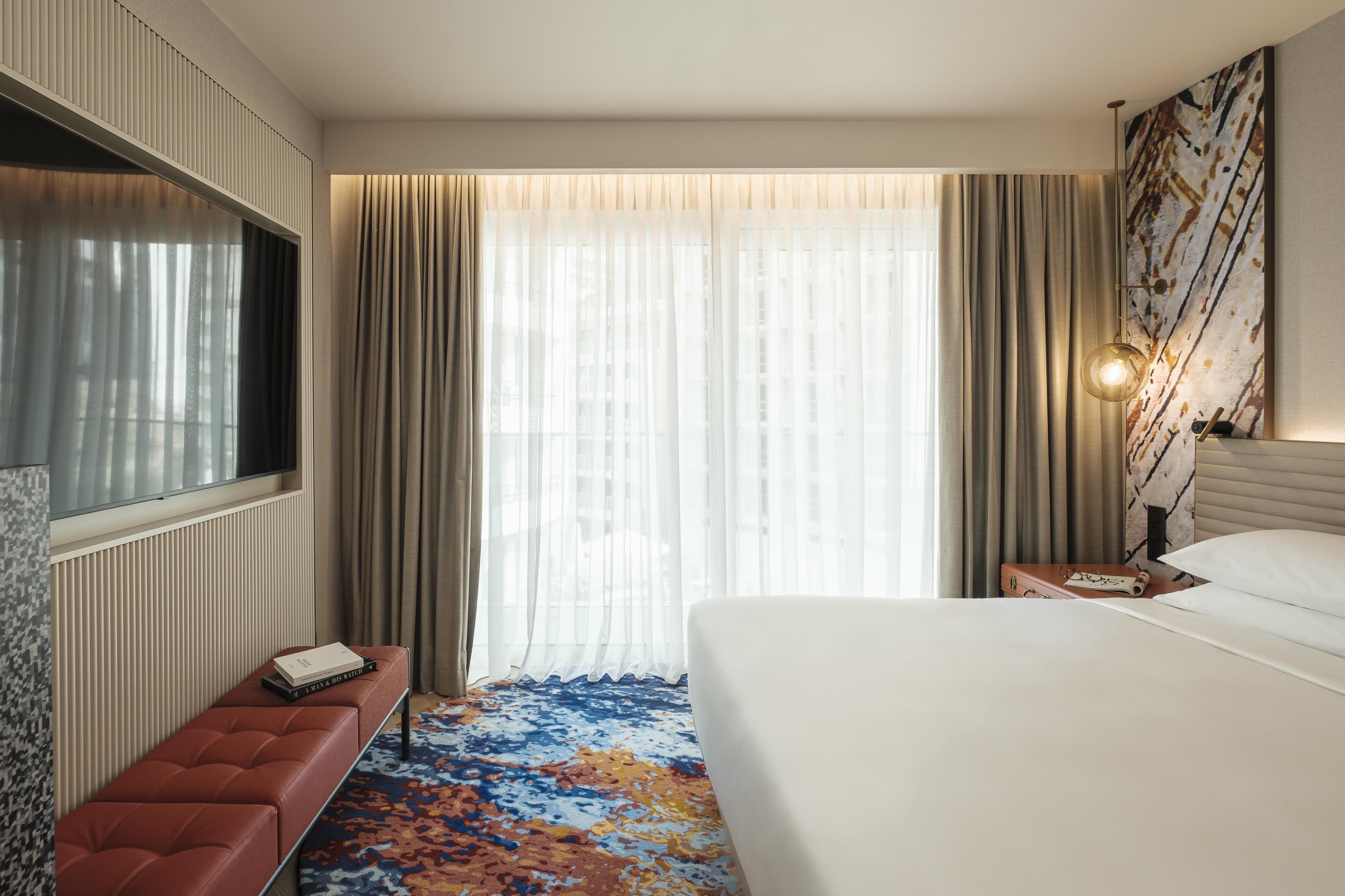 Une chambre d’hôtel avec un lit, une banquette, une télévision, une table de chevet et une grande fenêtre avec des rideaux.