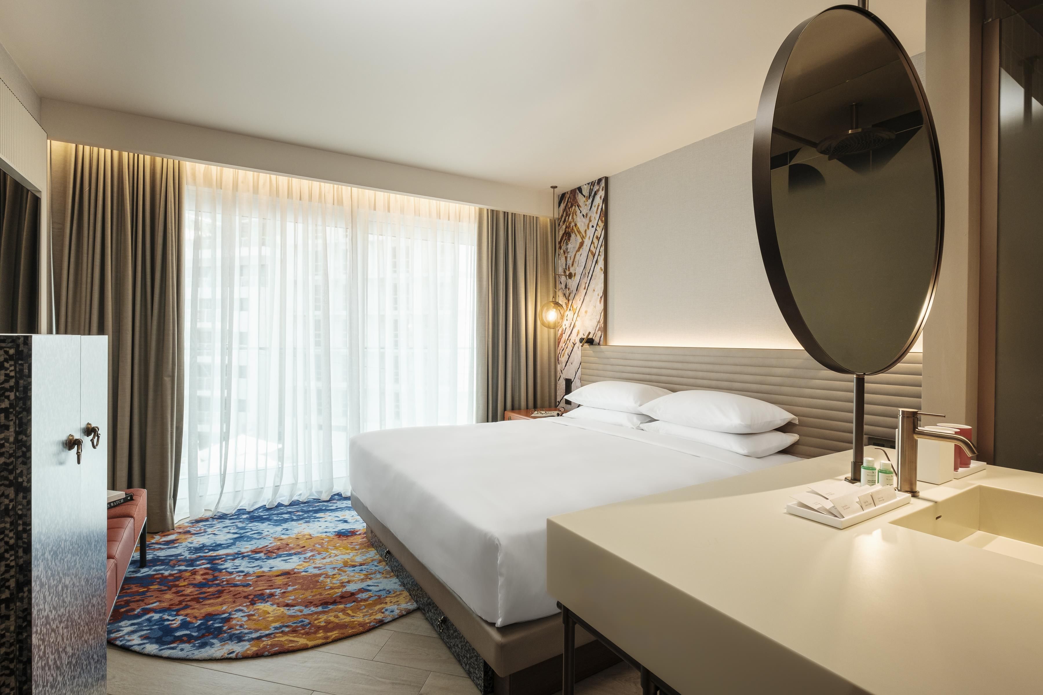 A hotel room with a bed, a bedside table, a mirror, a bathroom with a sink, and a window with curtains.