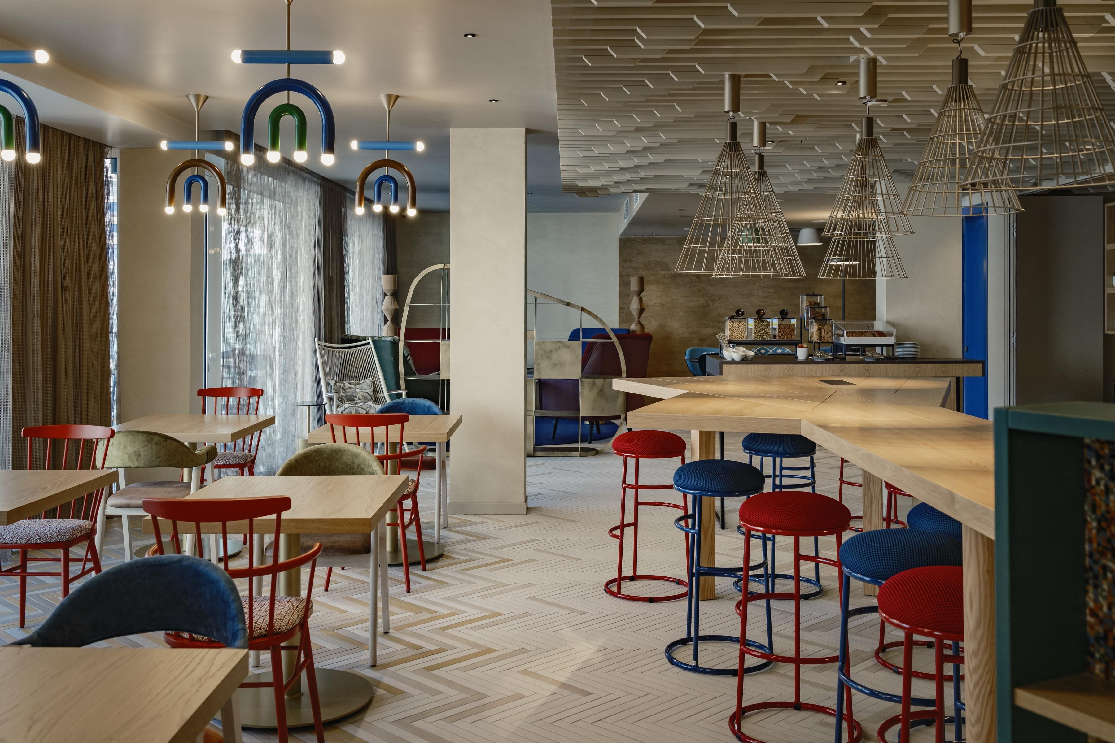 A modern restaurant interior with wooden tables, red and blue bar stools, and unique hanging light fixtures.