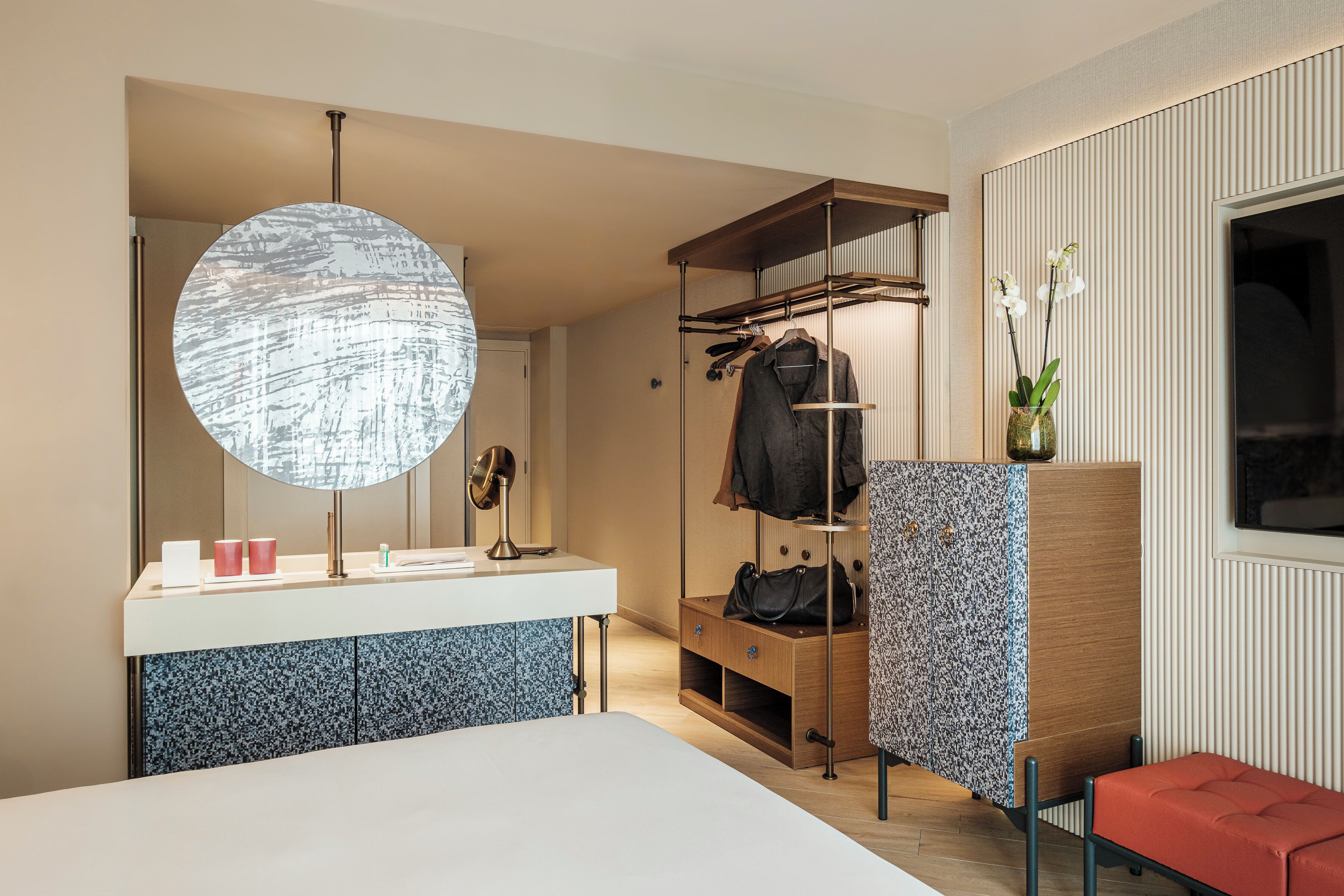 Una habitación de hotel moderna con un espejo grande, un espejo redondo, un tocador con lavabo, un perchero de madera, un banco y una ventana con vistas.
