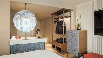 Una habitación de hotel moderna con un espejo grande, un espejo redondo, un tocador con lavabo, un perchero de madera, un banco y una ventana con vistas.
