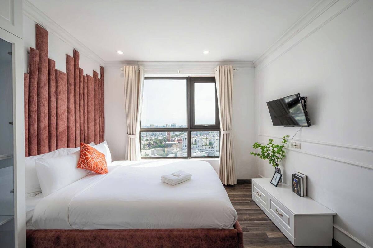A hotel room with a large bed, a TV mounted on the wall, a window with a city view, and a small plant on the side table.