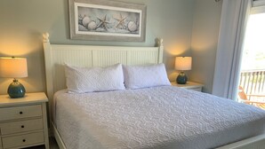 Una cama con cabecera blanca, dos mesitas de noche con lámparas, un cuadro enmarcado sobre la cama y vistas al exterior a través de una ventana.