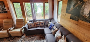 Interior acolhedor de uma cabana com um sofá de canto, duas poltronas e uma grande janela com vista para a floresta.