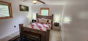 Un dormitorio con cama de madera, colcha, mesita de noche, lámpara de pared y ventana con vista a árboles.