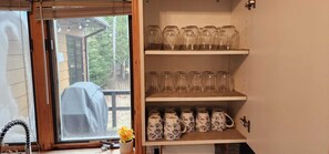 Une cuisine avec une fenêtre, une étagère où sont rangés des verres transparents et une rangée de tasses décorées.