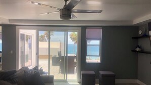 Uma sala de estar moderna com um ventilador de teto, um sofá, uma mesa de centro e vista para o oceano através de grandes portas de vidro deslizantes.