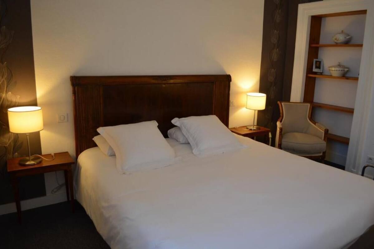 Een net opgemaakt bed met witte lakens en kussens, met aan beide zijden nachtkastjes met lampen, in een kamer met een houten hoofdeinde en een stoel met een plank tegen de muur.