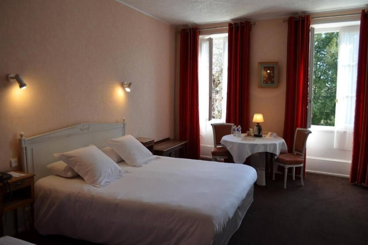 Een hotelkamer met een groot bed, twee nachtkastjes, een kleintje tafeltje met een stoel, en een raam met gordijnen.