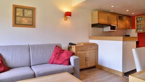 Ein kompakter Wohnraum mit grauem Sofa, einem kleinen Holztisch, einem roten Kissen, einer Küchenzeile mit Holzschränken und einer wandmontierten Leuchte.