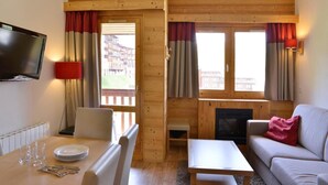 Ein gemütliches Zimmer mit Holzausstattung, einem Kamin, einem Sofa, einem Esstisch mit Besteck und einem an der Wand befestigten Fernseher.