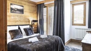 Un dormitorio con cabecera de madera, cama con ropa oscura, mesita de noche con lámpara, ventana con cortinas y vistas a las montañas.