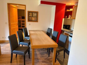 Uma sala de jantar com mesa e cadeiras de madeira, uma parede vermelha e uma cozinha ao fundo.