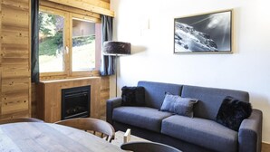 A cozy room with a wooden interior, a grey sofa, a fireplace, and a large window overlooking greenery. - 7th floor, view ski slopes, swimming pool, balcony, tv, ski locker, 41m², La Plagne (La Plagne)