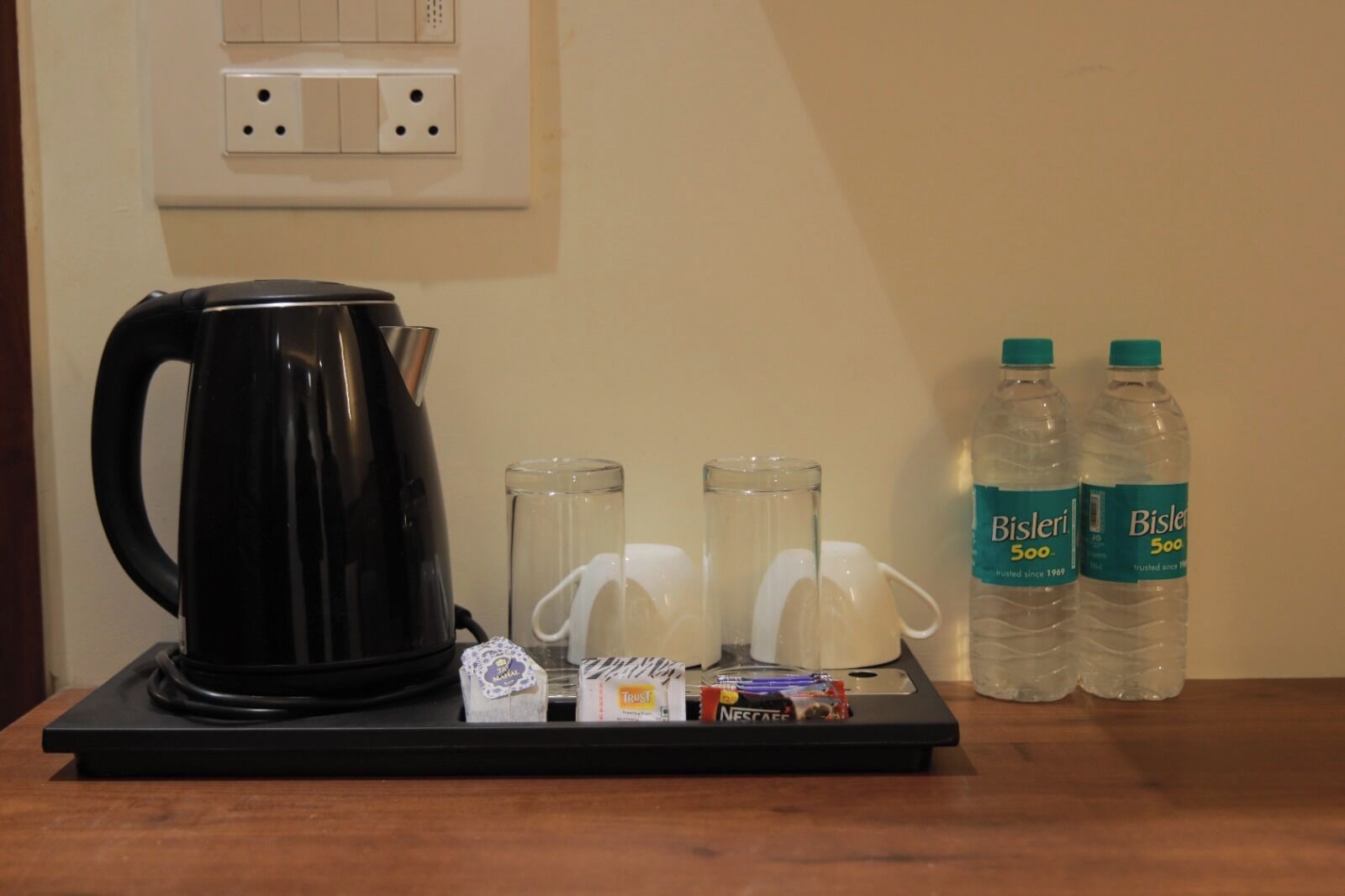 Una mesa con un hervidor eléctrico negro, dos vasos de vidrio, una pequeña bolsita de café Nespresso, un vaso de agua y dos botellas de agua Bisleri de 500 ml.