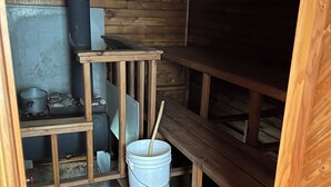 Een houten blokhut van binnen met een houten bank, een metalen emmer met een houten lepel en een open haard.