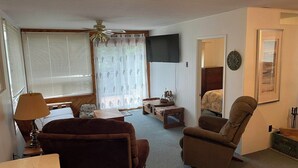 Een woonkamer met een bank, fauteuil, televisie en een raam met jaloezieën.