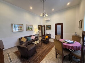 Un salon comprenant un canapé, des fauteuils, une table à manger recouverte d’une nappe rose et un lustre.