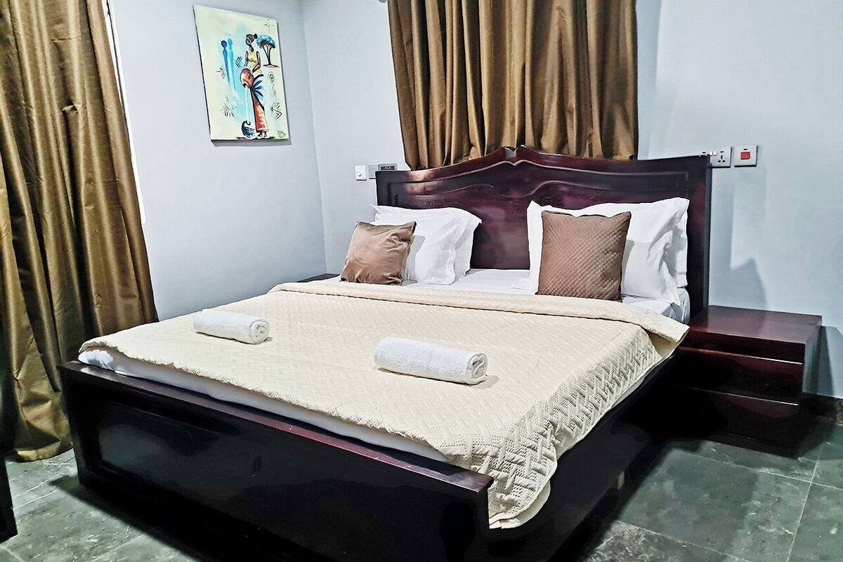 A neatly made bed with a beige blanket and two pillows, a wooden headboard, a nightstand with a lamp, and a painting on the wall.