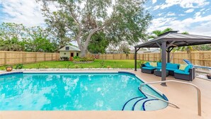 A swimming pool with a wooden deck, a covered seating area, and a large tree in the background.
