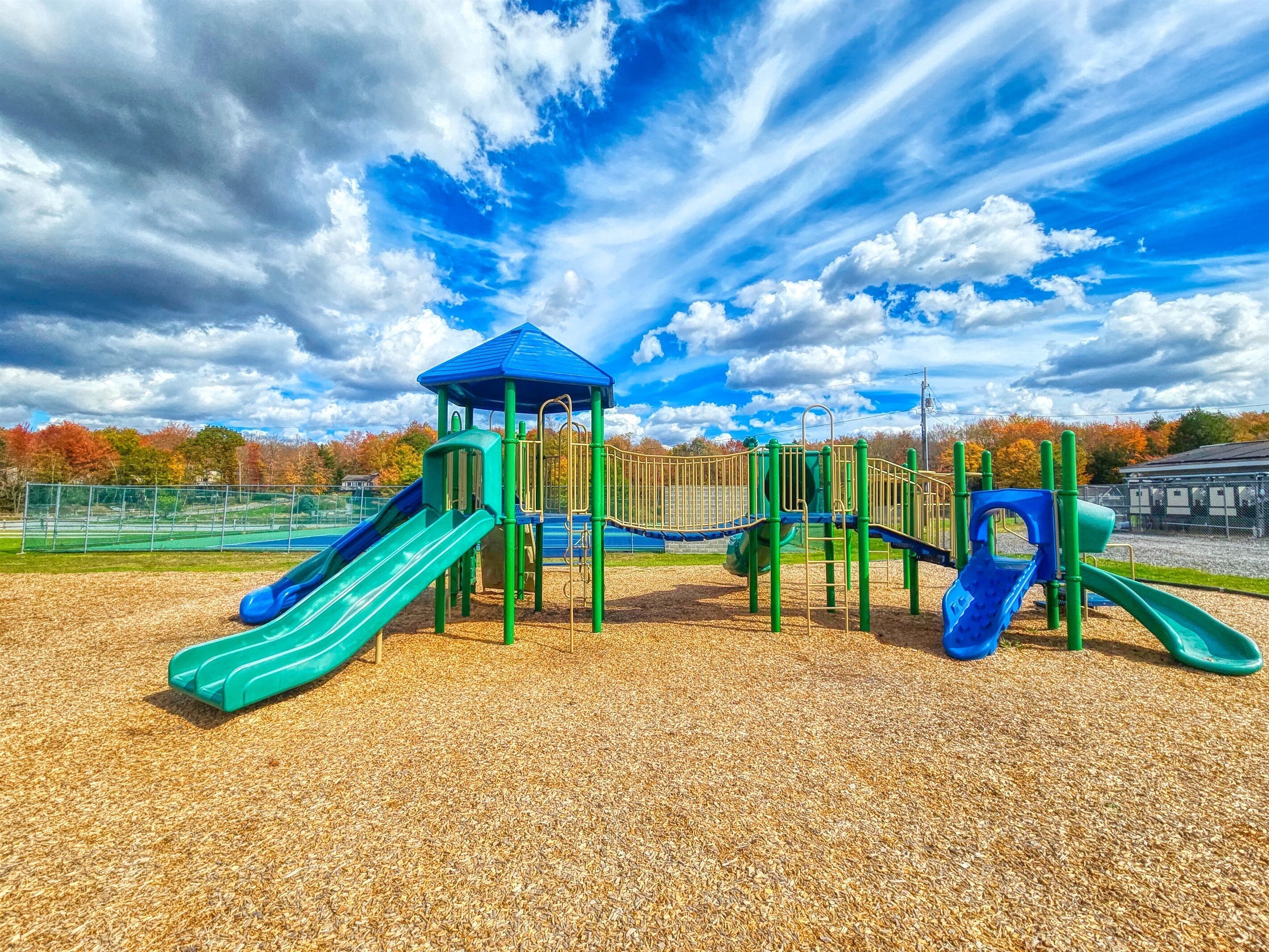 Un parque infantil con toboganes y estructuras para escalar, rodeado por una valla y árboles con follaje otoñal.