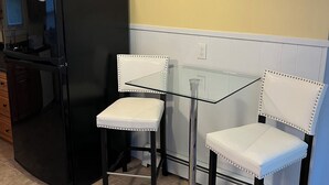 Une cuisine avec un réfrigérateur noir, deux chaises blanches à dossier clouté, une table en verre, des meubles de rangement en bois et une œuvre d’art murale sur le thème du « RELAX ».