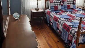Une chambre à coucher avec une commode en bois, un lit recouvert d’une courtepointe, une table de chevet avec une lampe et une horloge murale.