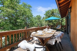 An outdoor deck with a wooden table set for dining, surrounded by white chairs, and a blue umbrella providing shade. - Modern Pine -  Walk to Bear Mtn, Close to Village, Mountain Top Views (Big Bear Lake)