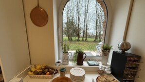 A kitchen counter with a fruit bowl, plates, a cake, and a pie, with a view of trees and a grill outside. - Aux Ecuries du Château  (Contremoulins)