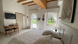 A bedroom with a large bed, wooden ceiling beams, a desk with chairs, and a TV on the wall. - Aux Ecuries du Château  (Contremoulins)