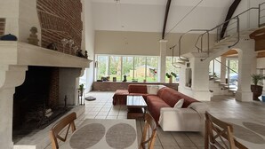A spacious living area with a fireplace, a sofa set, a dining table with chairs, and a view of the outdoors through large windows. - Aux Ecuries du Château  (Contremoulins)