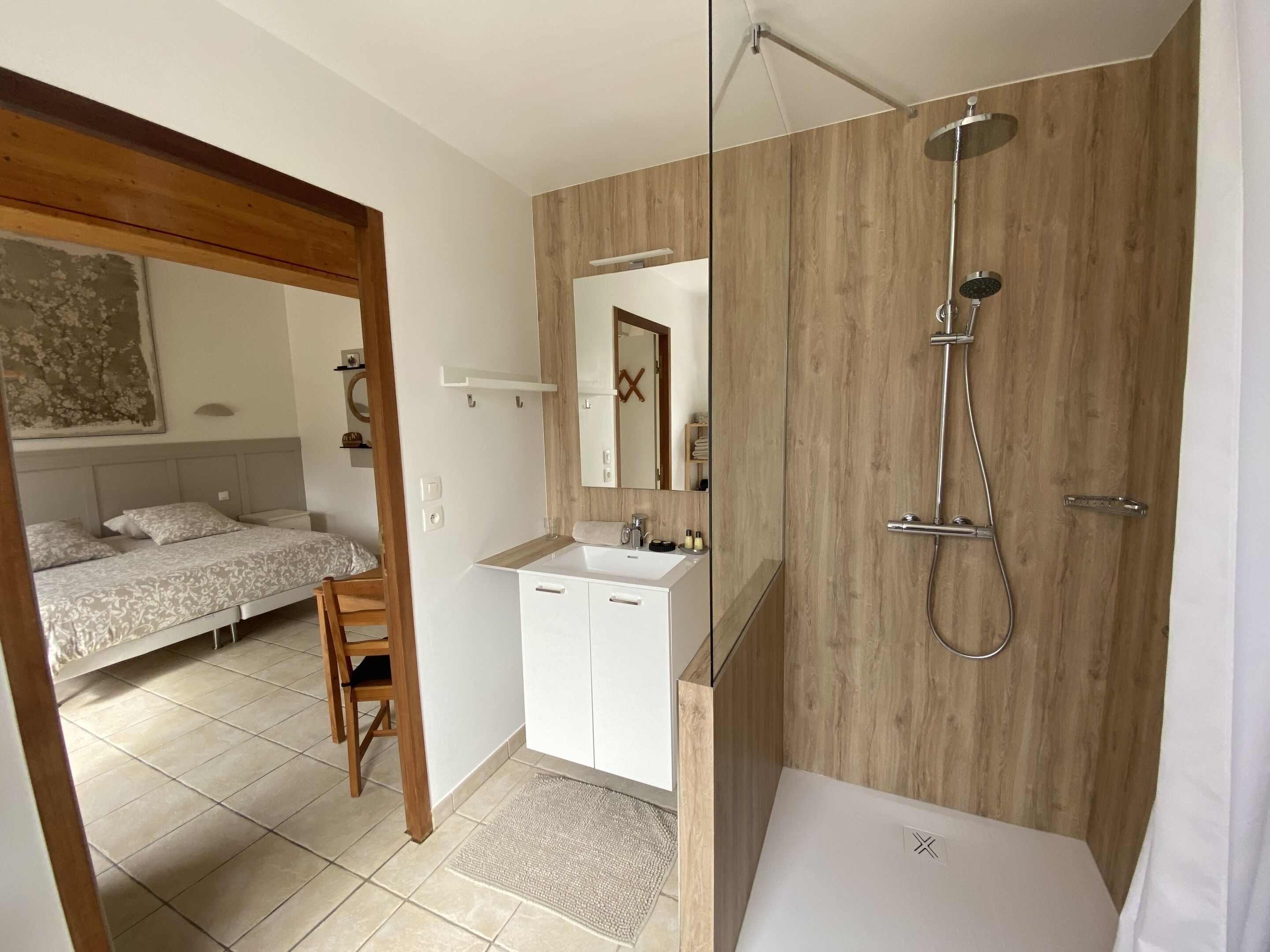 A modern bathroom with a shower, a sink, and a mirror. There is a bed and a chair in the adjacent room.