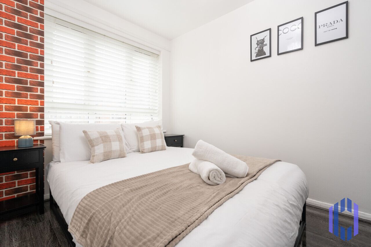 Een hotelkamer met een bed, een nachtkastje, een lamp, een raam met jaloezieën en drie ingelijste foto's aan de muur.