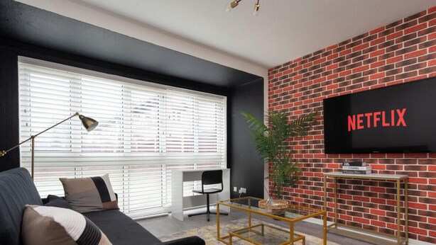 A modern living room with a black sofa, a coffee table, a TV mounted on a brick wall, and a desk with a chair. - Haven Noir by CEFAS - Pool Table - City & Man Utd (Greater Manchester)