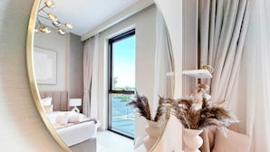 A modern hotel room with a large round mirror, a bed with white linens, a bedside table with a vase of dried plants, and a view of the outdoors through a large window. - Elegant 1B Apt&balcony in the Heart of Dubai Creek (Dubai)