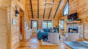 A log cabin interior with a blue sofa, stone fireplace, wooden floors, and a mounted deer head. - SPECIAL -  Make Your Labor Day a Mountain Getaway. Hot Tub, Fire Pit, Games (Mineral Bluff)