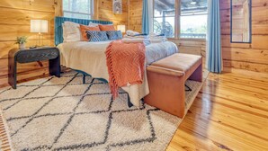A wooden cabin bedroom with a bed, bedside table, lamp, and a view of the outdoors through a window. - SPECIAL -  Make Your Labor Day a Mountain Getaway. Hot Tub, Fire Pit, Games (Mineral Bluff)