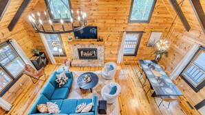 A cozy cabin interior with a fireplace, wooden floors, a sofa, a dining table set for four, and large windows. - SPECIAL -  Make Your Labor Day a Mountain Getaway. Hot Tub, Fire Pit, Games (Mineral Bluff)